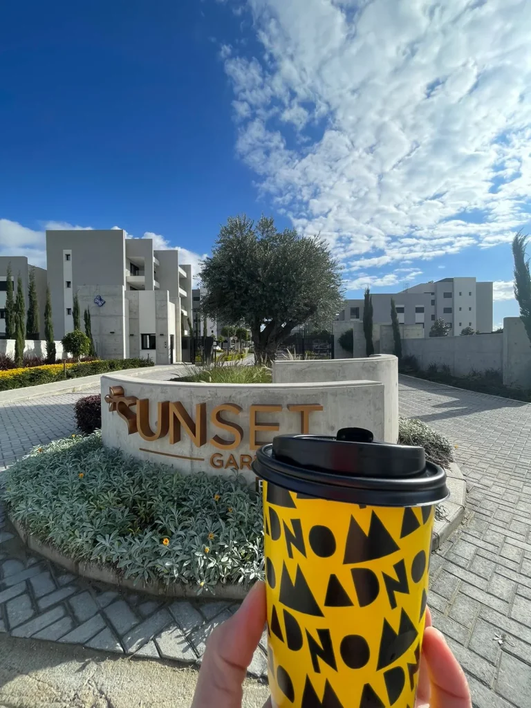 black and yellow coffee cup, sunset gardens entrance, olive tree, buildings, blue sky