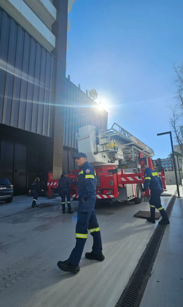 Firefighters in uniform conduct a safety drill outside a modern building, using a fire truck with an extended ladder under a bright sun.